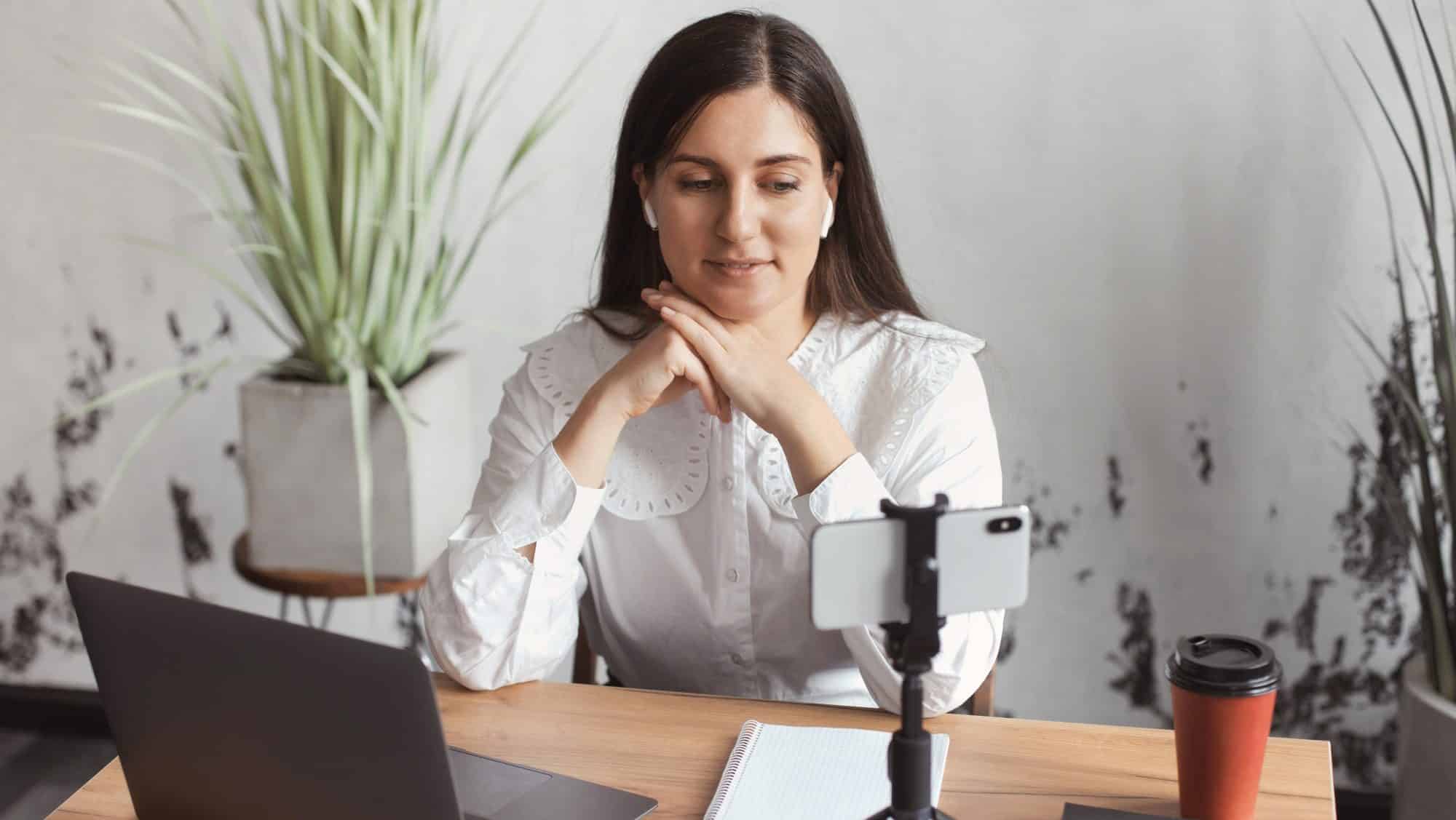 Business woman in wireless headphones working on laptop and listening online webinar on smartphone